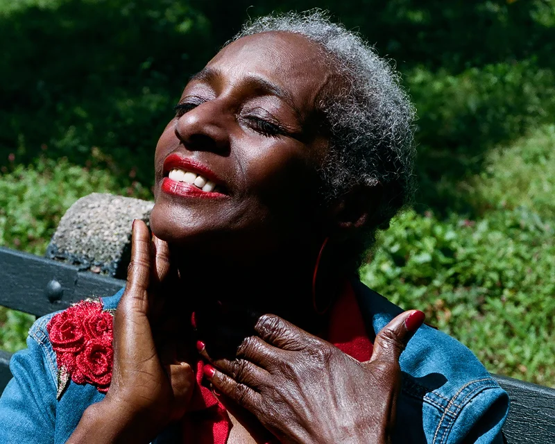 This image shows a portrait of a Black older person outdoors with their head tilted back and eyes closed. The person is wearing red lipstick and large red earrings.