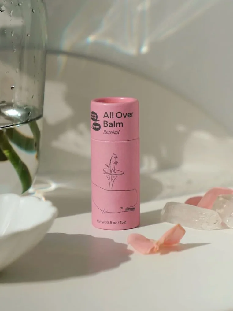 A pink tube of balm stands on a white table top with crystals and flower petals strewn next to it.