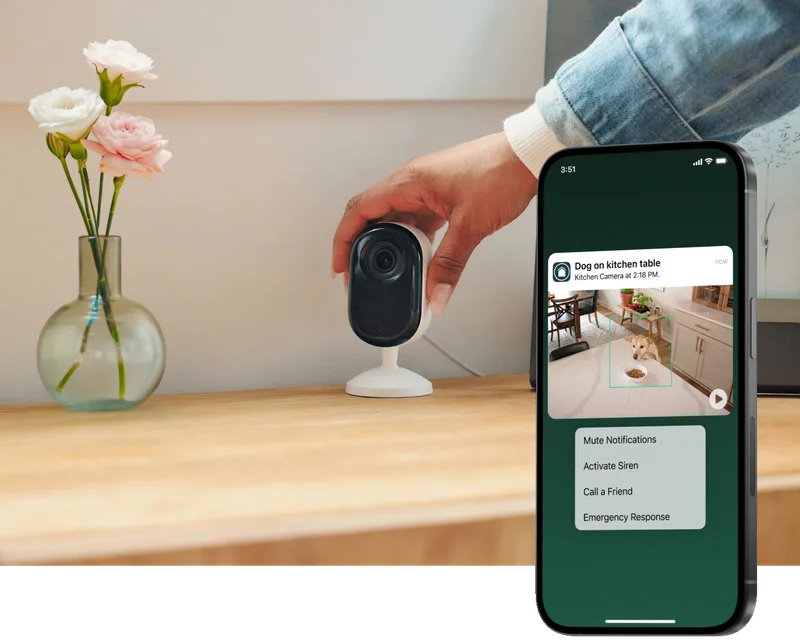 Phone displaying a simulated screen image of an Arlo Secure notification with animated preview. Background image depicts someone setting up an Arlo Essential Indoor Camera next to flowers on a table.