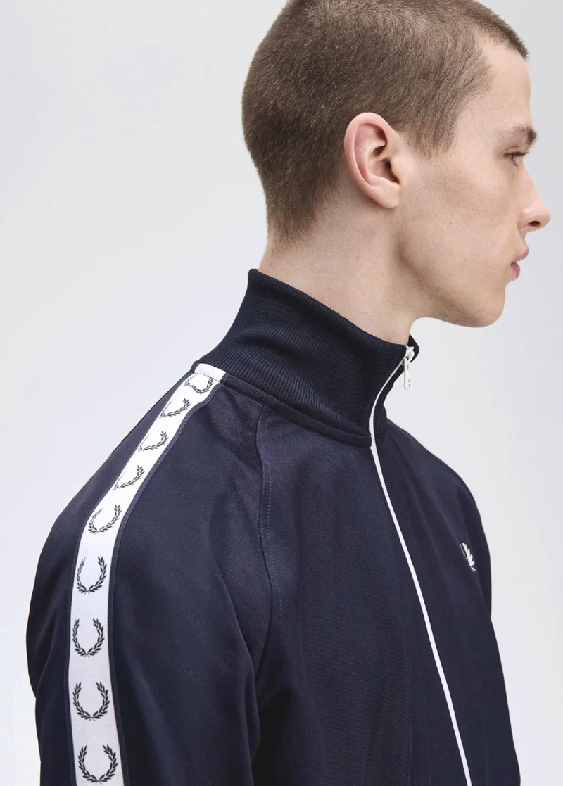 white male model with buzzcut wears a navy track jacket with contrasting white taping down the shoulders and arms