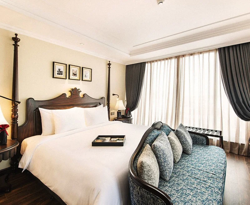 Image description: Hotel room with large bed and stately wooden bed frame. There is a blue chaise at the end of the bed. Sheer curtains are drawn on the window.