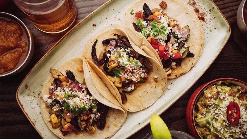 Image description: Overhead view of three tacos on a plate with drinks and guacamole