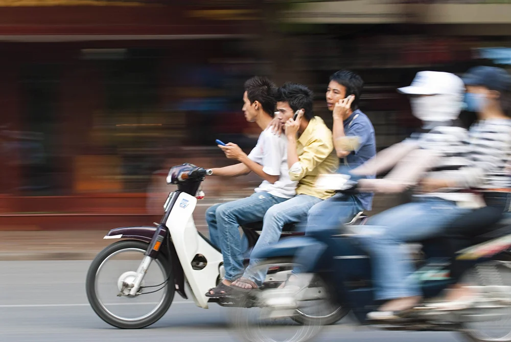 Three young men using their phones while riding on a scooter.