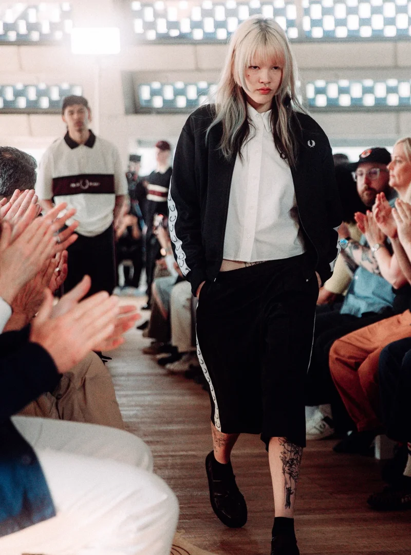 White female model with blonde hair with a layered fringe walks the runway wearing a white shirt, a classic black track jacket with white laurel wreath taping, and a midi black skirt with white laurel wreath taping