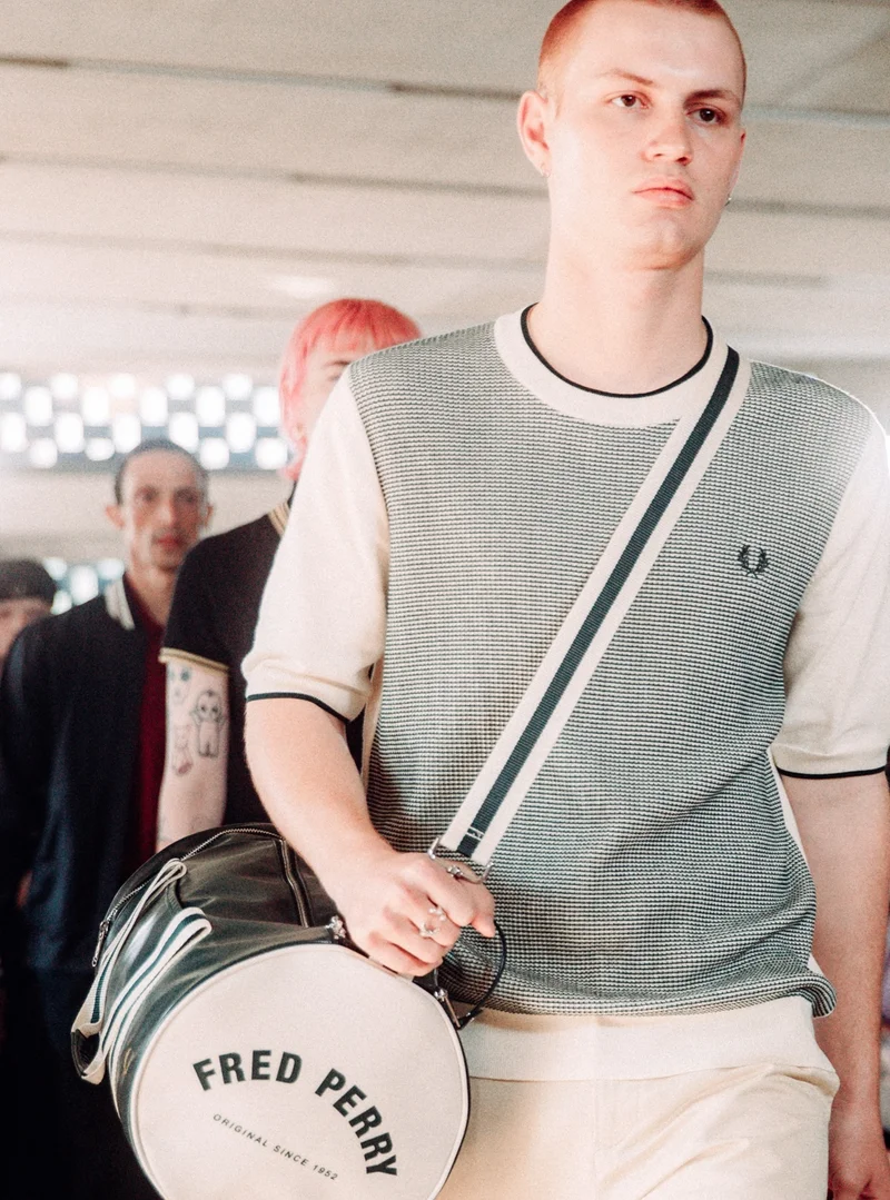 a line of models wall the runway. At the front is a white male model with a ginger buzzcut, wearing a Tonic Waffle knitted T-shirt in ecru and black, with a classic black and white barrel bag across his body