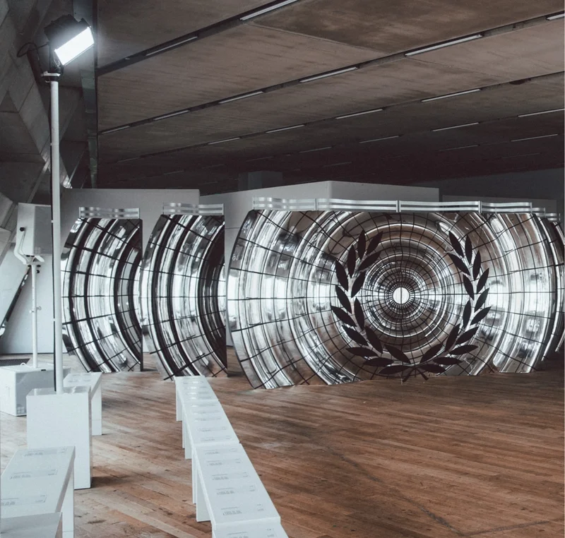 this image shows the set of the runway, one floor of the Tate Modern, with wood panelled floors, white benches, a industrial ceiling and huge art installations reminiscent of speakers with a laurel wreath at the centre