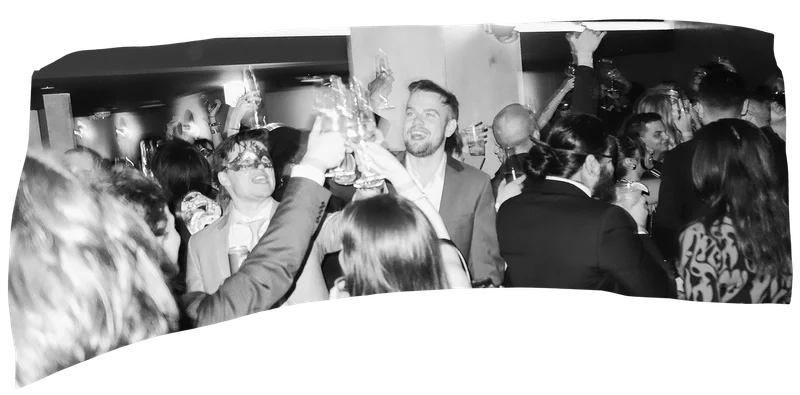 Black and white photo of a lively party with guests raising their glasses in a toast.