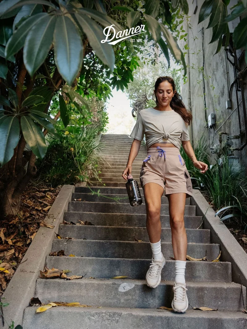 A woman walks down outdoor concrete steps surrounded by greenery, wearing the Danner Women's Trail Trainer in Sand/Semolina, beige shorts, and a knotted beige t-shirt.