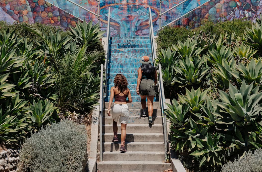 Two women walk up colorful outdoor steps surrounded by lush greenery, both wearing the Danner Women's Trail Trainer
