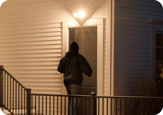 Burglar near the front door of a home with an Arlo Wired Floodlight lighting up the area surrounding.