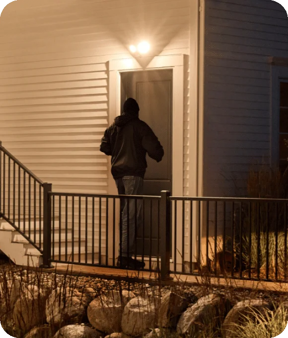 Burglar near the front door of a home with an Arlo Wired Floodlight lighting up the area surrounding.