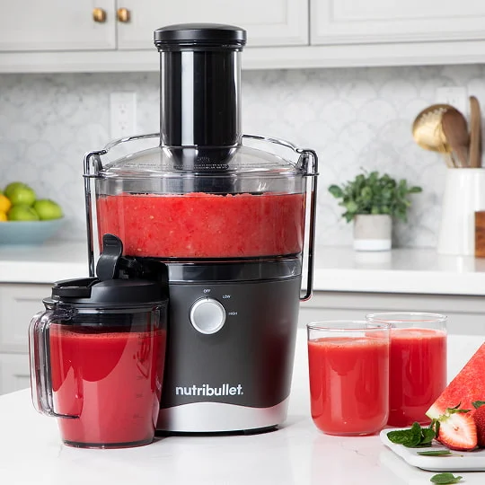 A nutribullet Juicer™ with red juice sits on a kitchen counter. Nearby are a pitcher, two glasses, and a cutting board with fruit.