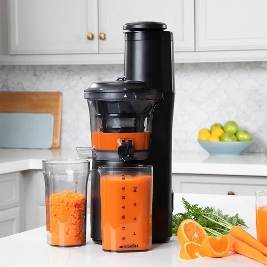 A black nutribullet® Slow Juicer with orange juice sits on a kitchen counter. In front, two pitchers hold juice and pulp. Carrots, and an orange lie nearby, with a tiled backdrop.