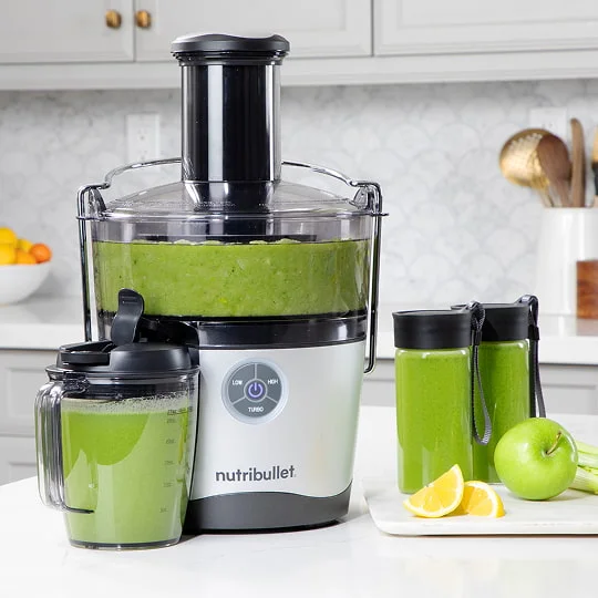 A nutribullet Juicer Pro™ with green juice sits on a kitchen counter. Nearby are a pitcher, two bottles, and a plate with an apple and lemon wedges.