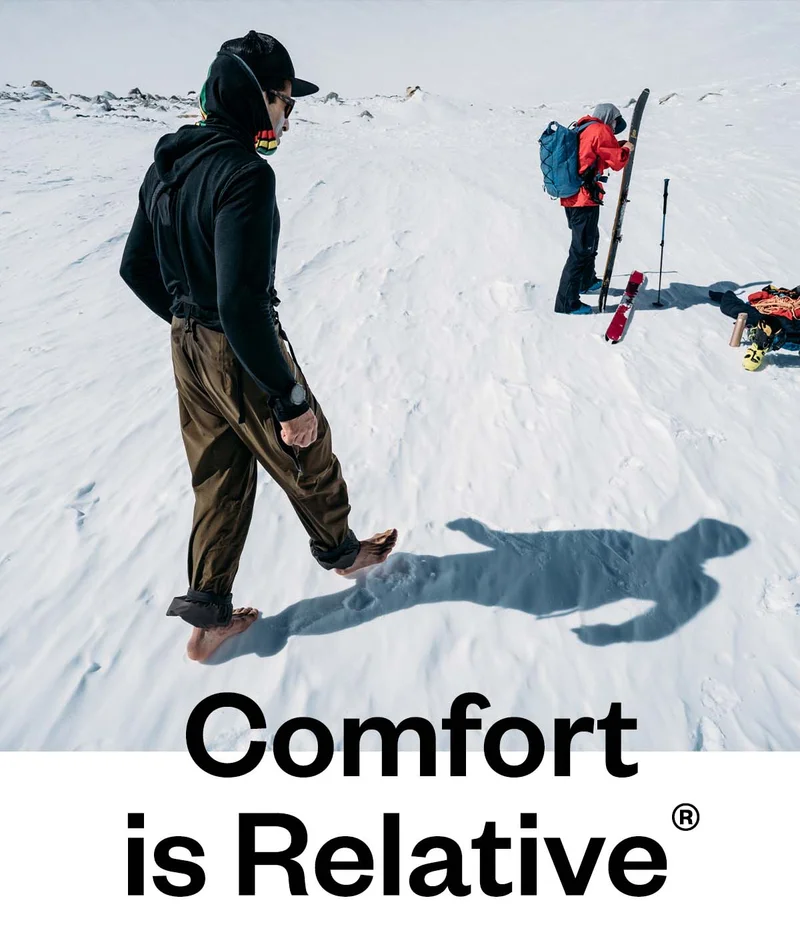 Comfort is relative. A person walks barefoot in the snow toward their skiing partner and a pile of gear.
