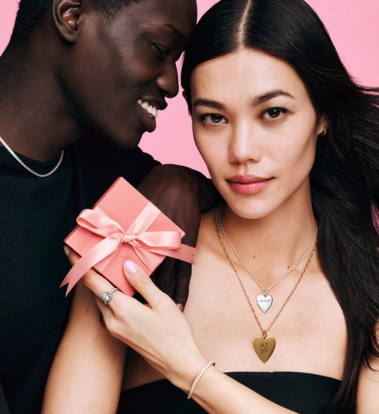 A woman and her male partner in an embrace. The woman is holding a pink gift box and wearing heart engraving charm necklaces.