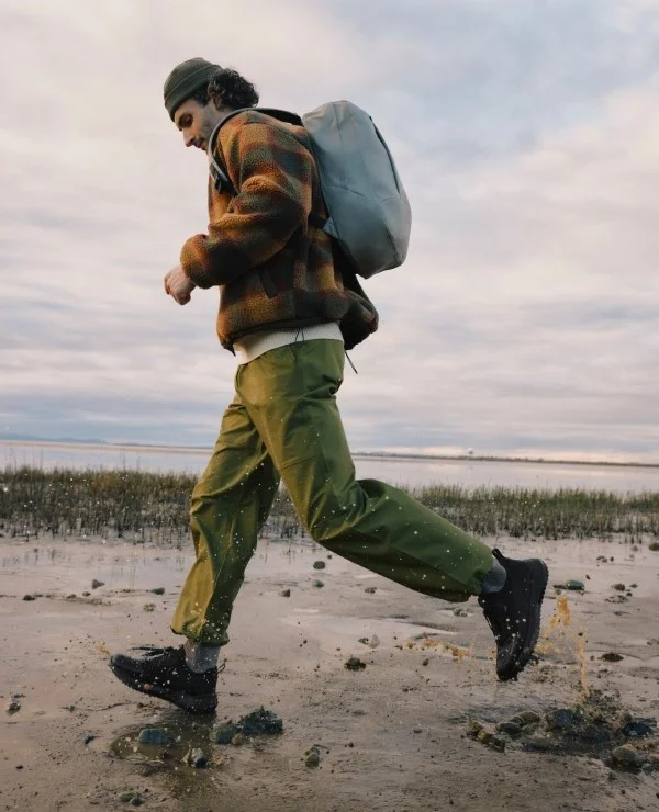 Graphic image of a man running in a muddy water wearing a Stormburst Low Top in Black and carrying the new Astoria Backpack.