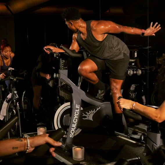 Photo of a person wearing black shorts and a black tank top on a SoulCycle stationary bike. They are wearing a Fitbit device on their wrist.
