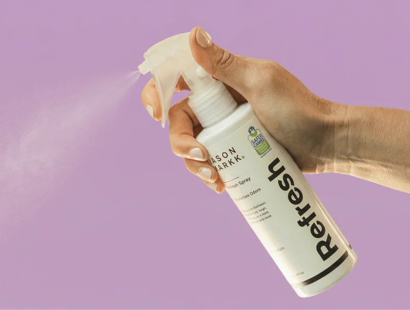 A medium shot shows a hand holding a white spray bottle with a clear nozzle, spraying a fine mist against a solid lavender background. The bottle has ...