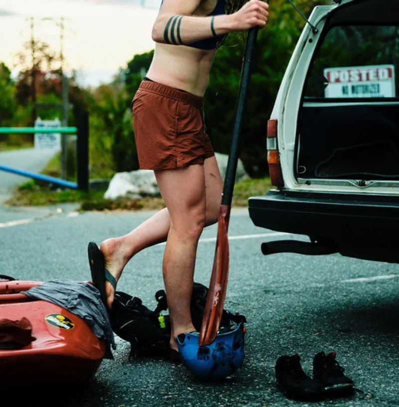 A person wearing wet shorts, a sun hoody and a puffy vest leans against an old Volvo with a kayaking helmet on the hood and a kayak in the foreground.
