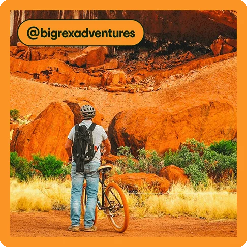Image description: A man with a bike helmet and backpack standing to the left of a bicycle in front of a red cliff, photo taken by @bigrexadventures