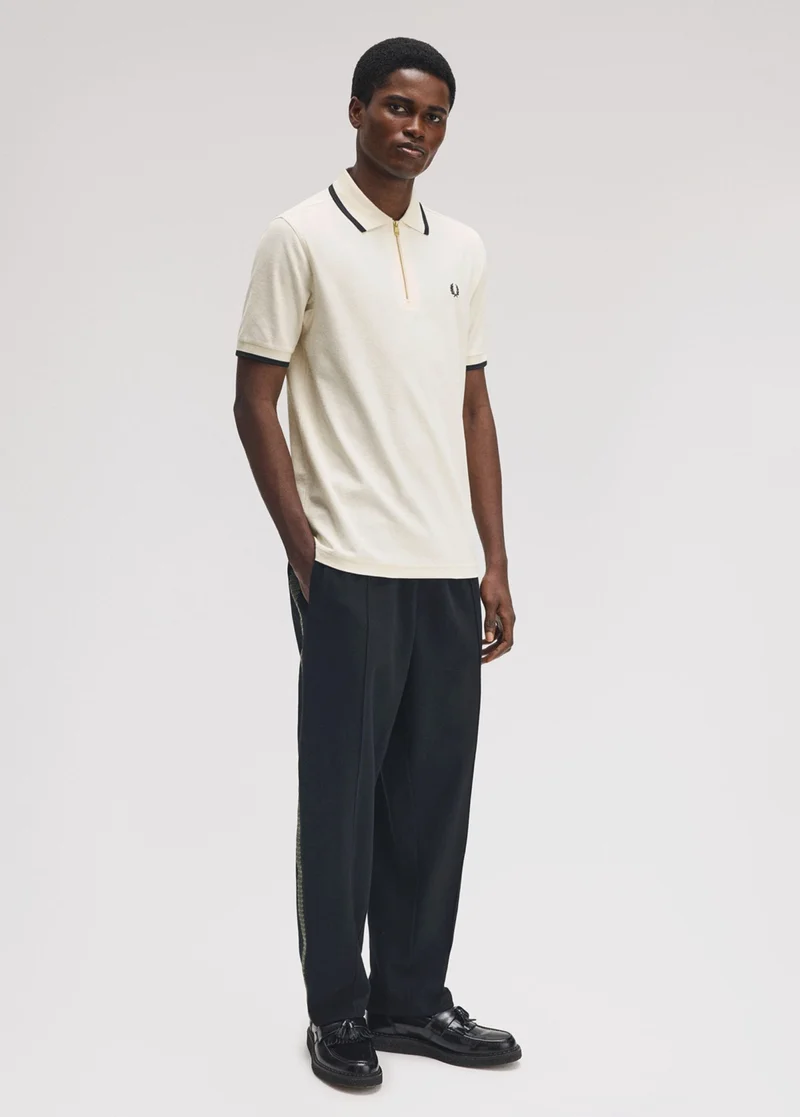 Black male model with short hair stands with a hand in his pocket wearing an ecru coloured crepe polo shirt with quarter zip fastening and navy single tipping