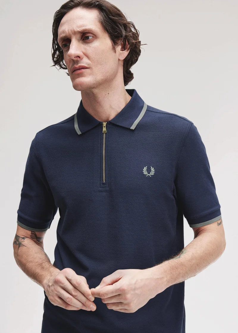 White male model with dark ear-length hair looks to the side wearing a navy coloured crepe polo shirt with quarter zip fastening and sage green single tipping