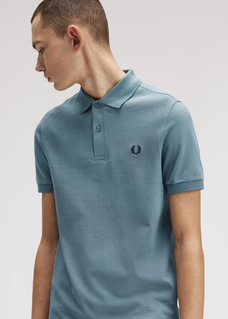 Young white male model with buzzcut is turned to the side and wears a dusty blue one-colour pique polo shirt