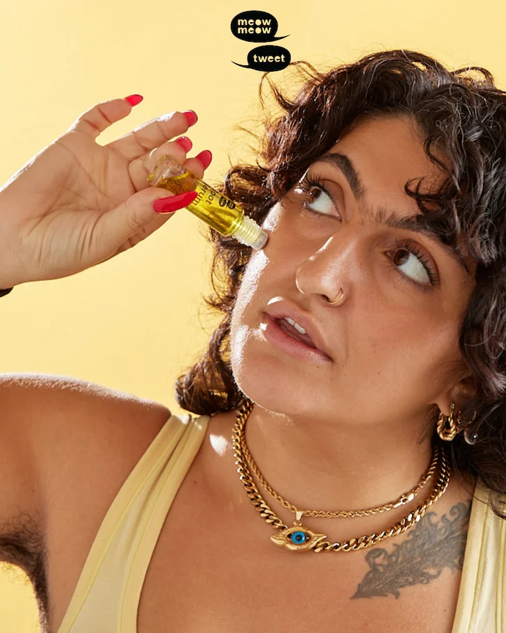 A tan person with dark brown curly hair and long red nails, tips their head back and applies a roller ball of spot serum to their cheek.