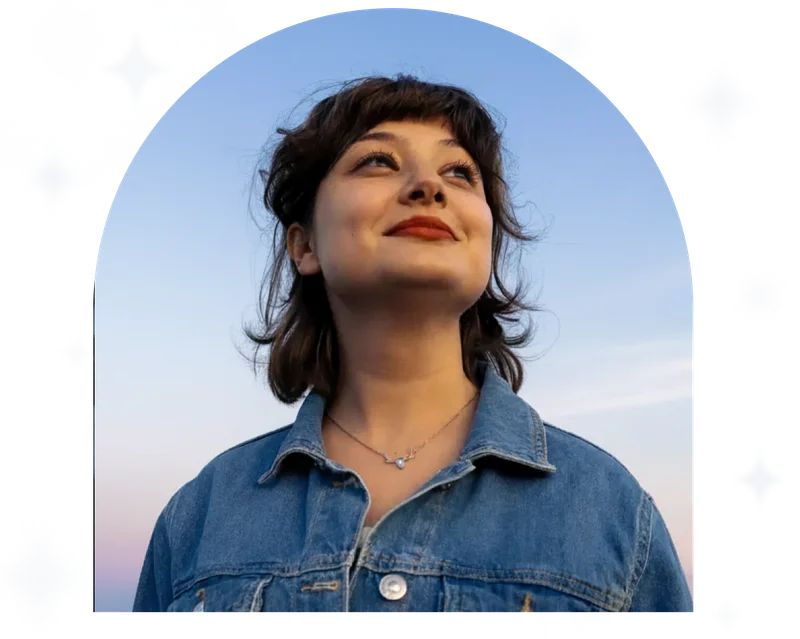 Photograph of a beautiful young woman wearing a denim jacket and smiling towards the sun, taken in the sweet pastel colors of the sunset.