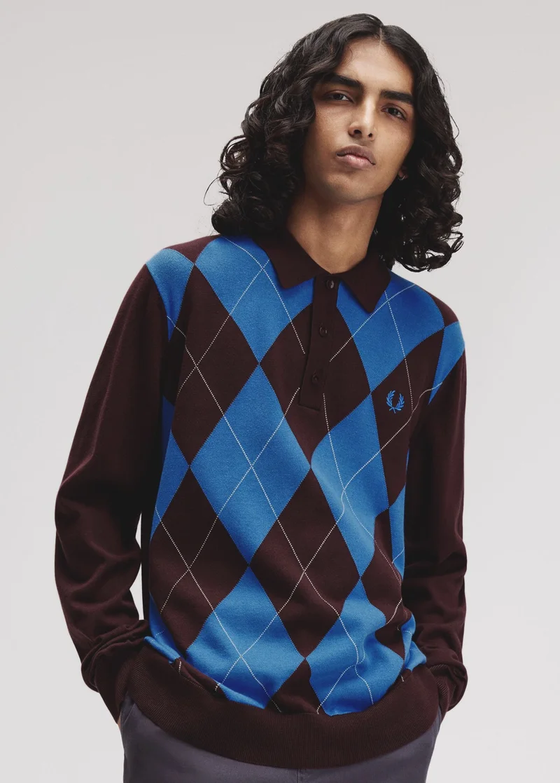 male model with brown skin and long luscious wavy hair stands with his hands in his pockets and wears a knitted long sleeved polo shirt with a bright blue and deep brown diamond argyle pattern