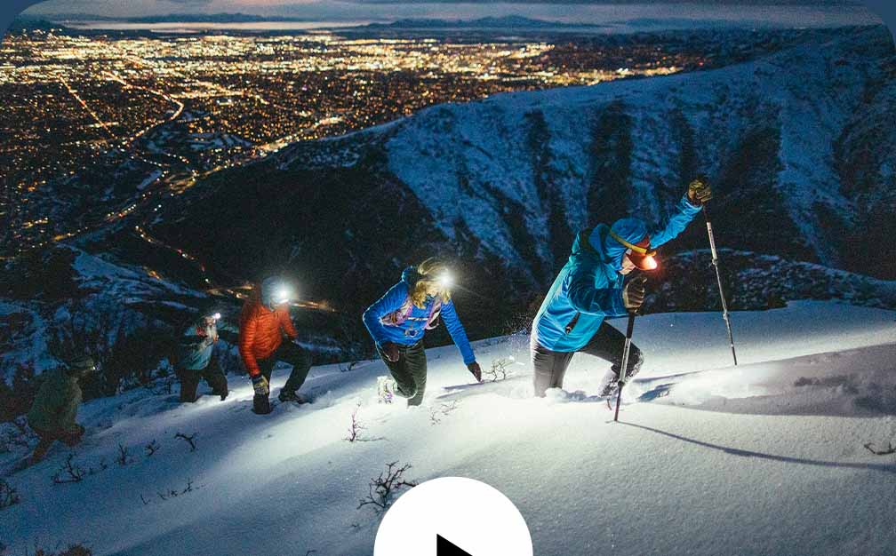 A group of people running up a snowy mountain in the dark with city lights peppering the background.