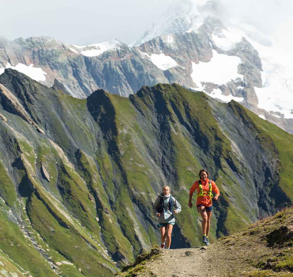 Two people running on a trail through snowy mountains.