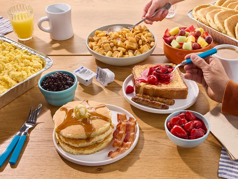 A full spread of IHOP Catering on a table, including tin trays filled with scrambled eggs and pancakes, a plate of pancakes and bacon, cups of fruit, and French Toast