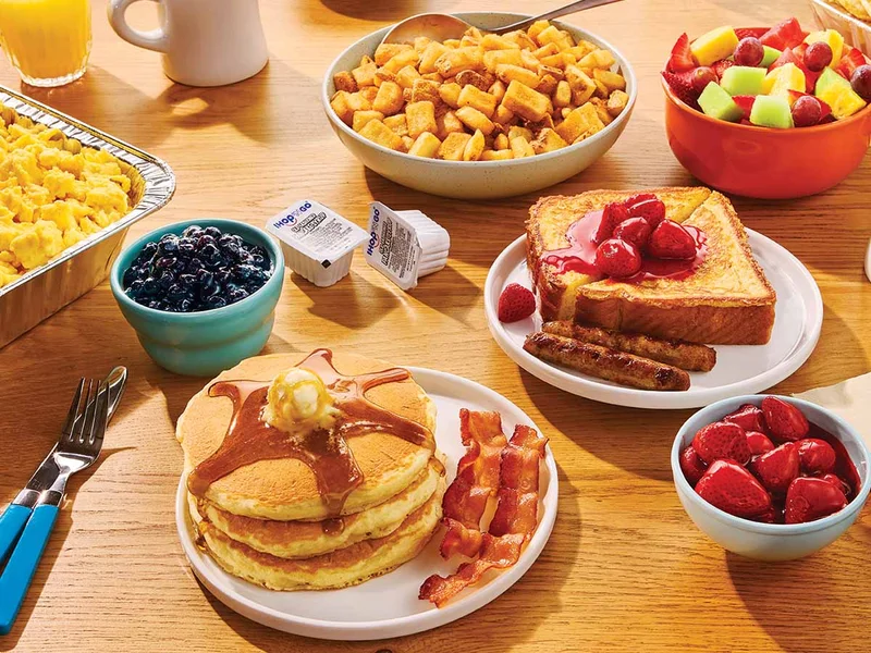 A spread featuring plates of IHOP Catering on a wooden table. Items include bowls of berries, plates of pancakes and french toast, and a tin of scrambled eggs.