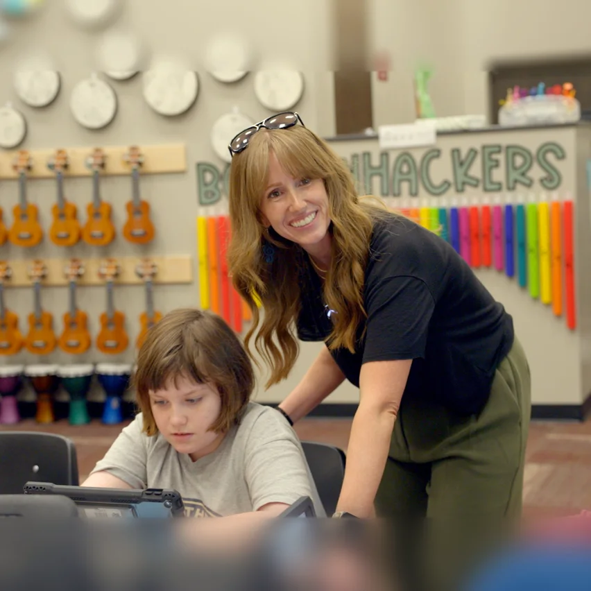 Teacher assisting a student with a tablet in a music classroom.