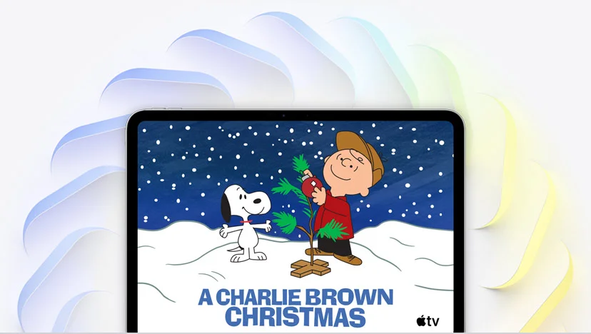 iPad Pro, 11-inch, silver color, on-screen Charlie Brown decorates small Christmas tree, Snoopy watches, snow falls, and Apple TV logo in bottom right corner, multiple angled squares with rounded corners in blue color, green color, and yellow color fan out from behind