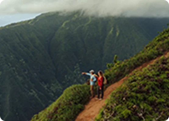 Two hikers standing on a narrow mountain trail overlooking deep green valleys and misty peaks.