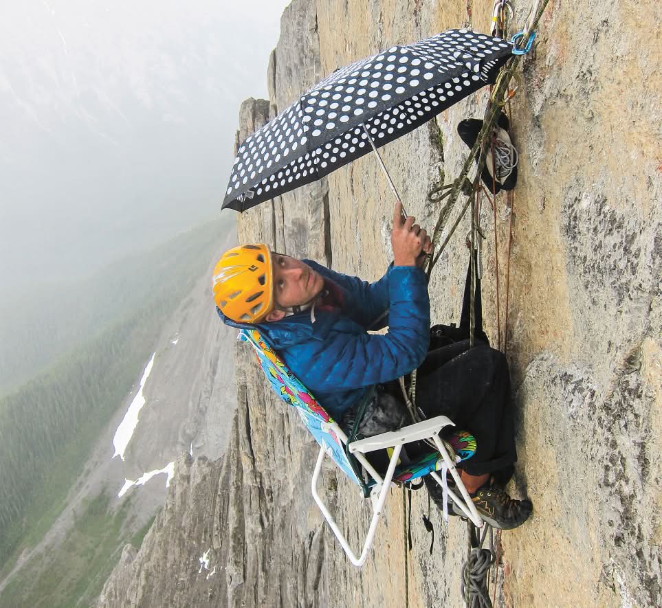 A climber holds an umbrella while sitting in a folding chair high on a rock wall.