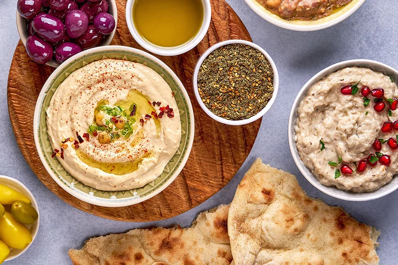 A festive spread featuring hummus topped with herbs and pomegranate seeds, surrounded by pita, small bowls of spices, grains, and grapes.