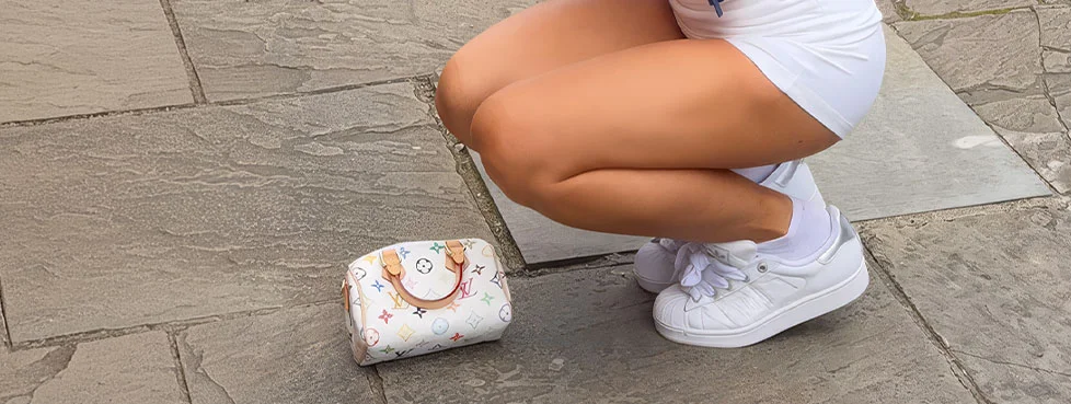 Person squatting with a white Louis Vuitton Monogram Multicolore Speedy bag, white shorts, and white sneakers on stone pavement.