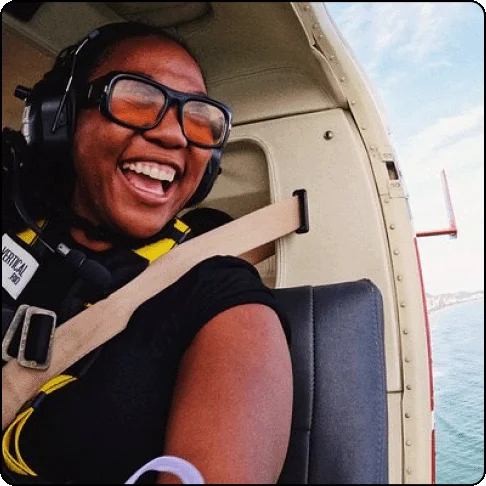 Image Description: Woman wearing tinted sunglasses and a headset smiles big as she looks out from the seat of a helicopter.