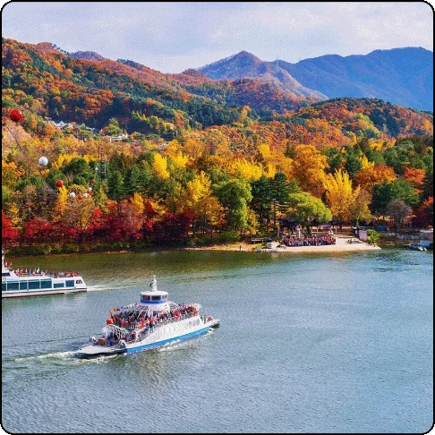 Image Description: There is a calm, large body of water with two boats in the foreground, with mountains and trees in the background showing a wide array of colors from green to orange and yellow.