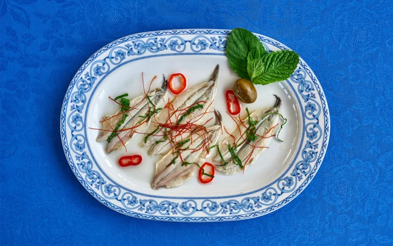 A dish of four marinated anchovies topped with sliced red peppers, an olive and a sprig of mint served at a restaurant on Stromboli Island, Italy.
