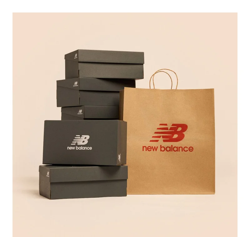 Black shoe boxes with a white New Balance logo stacked next to a brown paper shopping bag with a red New Balance logo.
