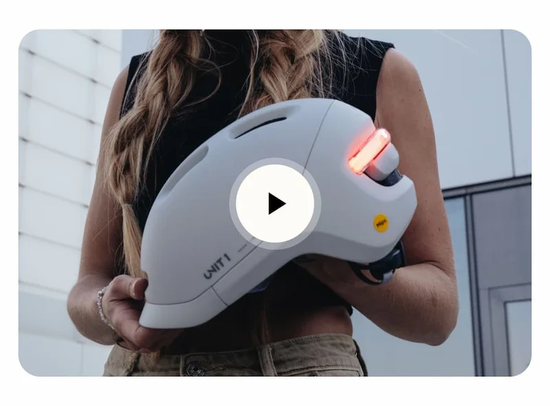 Person holding the UNIT 1 NEON Smart Helmet with rear light on and a play button overlayed on the image.