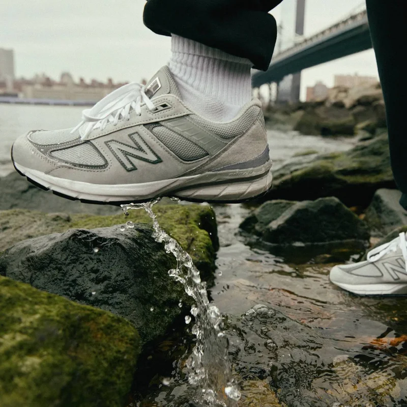 A medium shot captures a person wearing a grey New Balance sneaker with white laces and white socks, stepping over rocks in shallow water with a bridg...