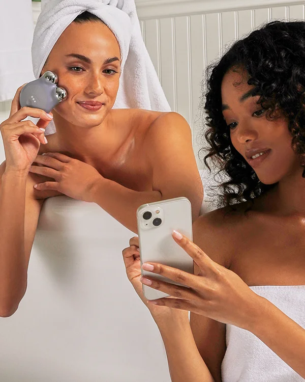 Blonde woman with hair slicked back holding along cheek contour with TRINITY+ Device, following along NuFACE Smart App on phone