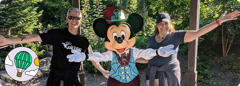 Image Description: Two people posing for a photo with Mickey Mouse. An Experience Explorer badge is on the bottom left corner of the image.