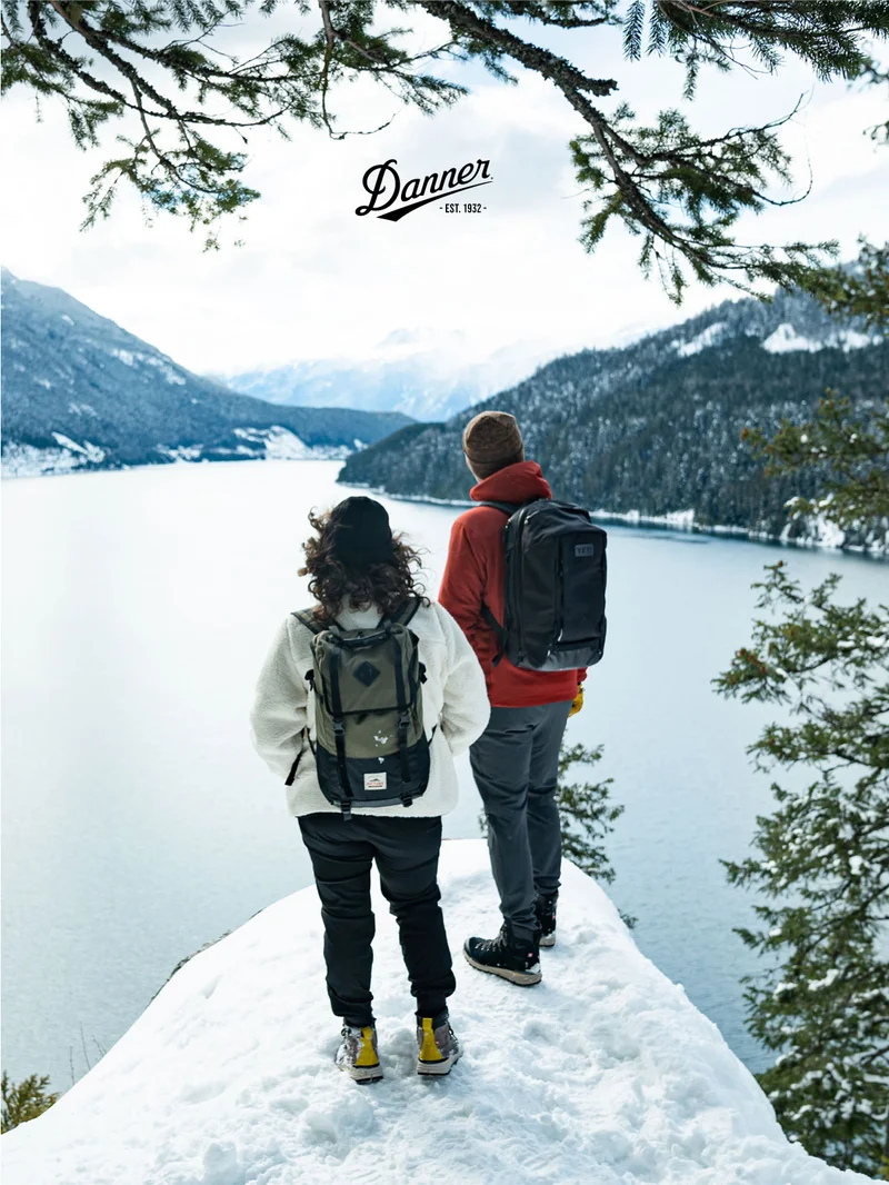 A man and a woman standing on a snowy overlook, gaze at a mountainous landscape across a lake. The Danner logo is overlaid at the top.